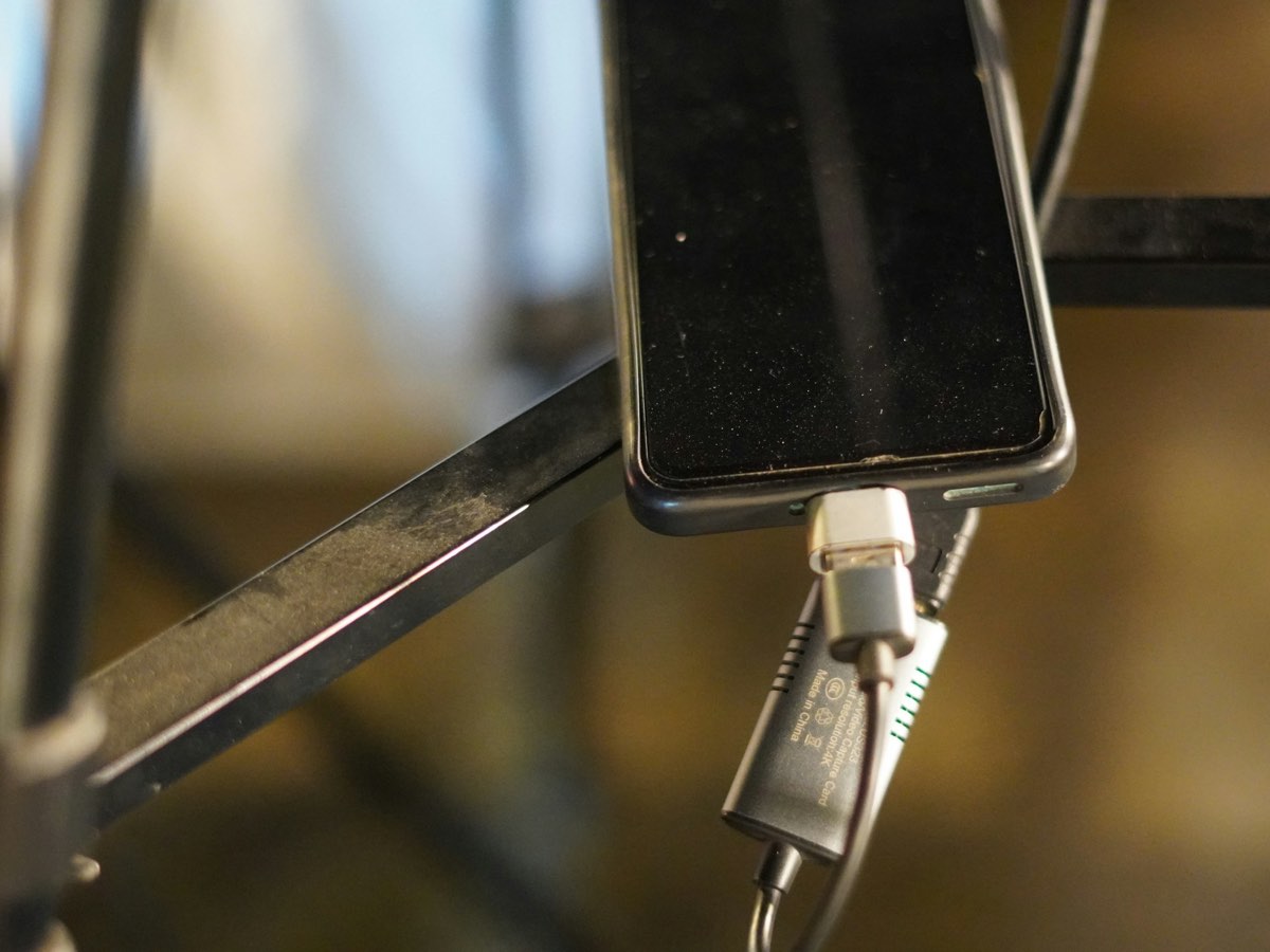 A phone on a metal stand with a silver adapter in its charging port, a full-size USB plug in the adapter and a small USB video device hanging from it on its cable.