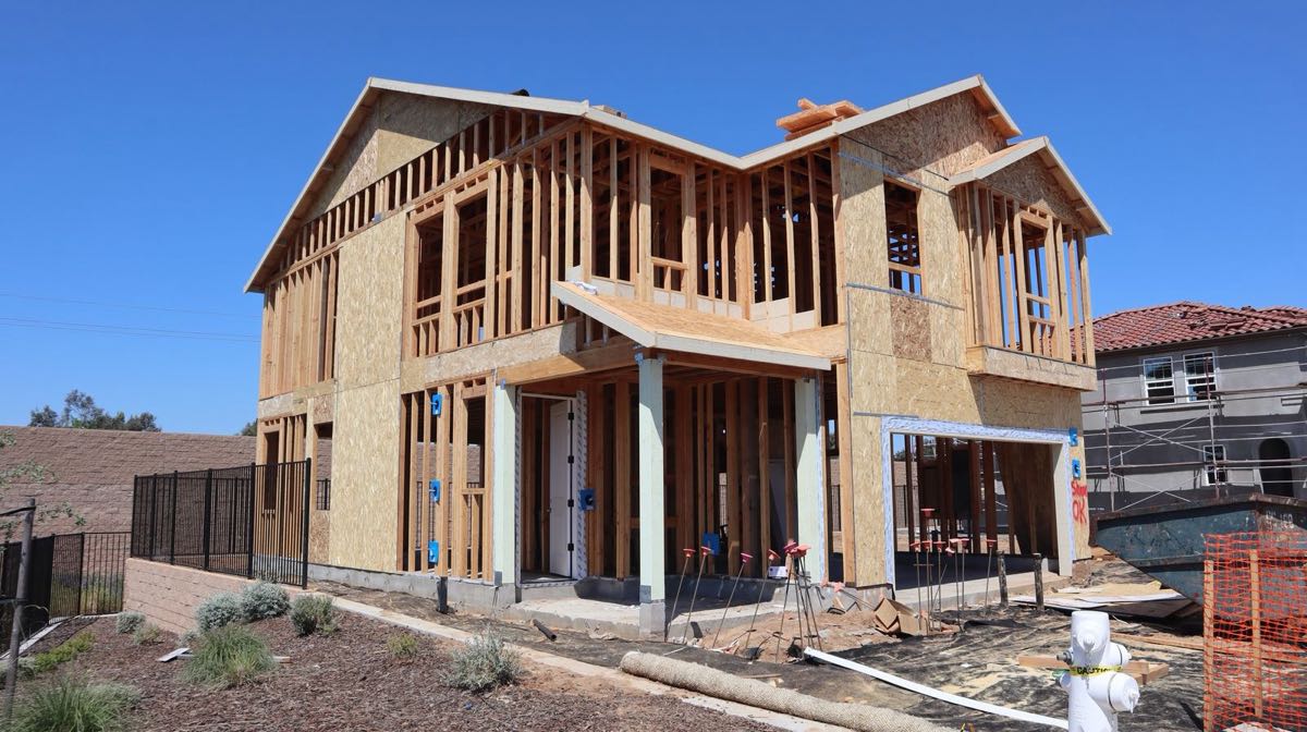 A two-story house at the framing stage on a sunny day: bare studs on the upper floor, sheathing on parts of the walls, an open front porch, electrical boxes on the studs and a dumpster at the side.