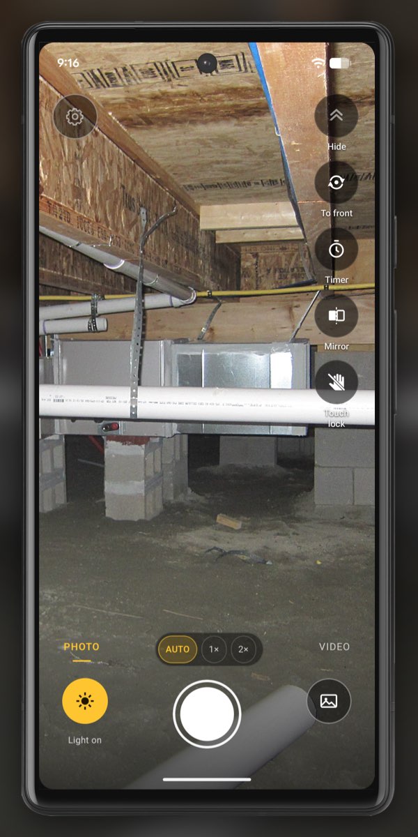 InspectCam on an Android phone looking into a crawl space with Light on lit: the white drain pipe, ductwork and block piers up close are bright, and the back of the crawl space behind the piers is already dark.