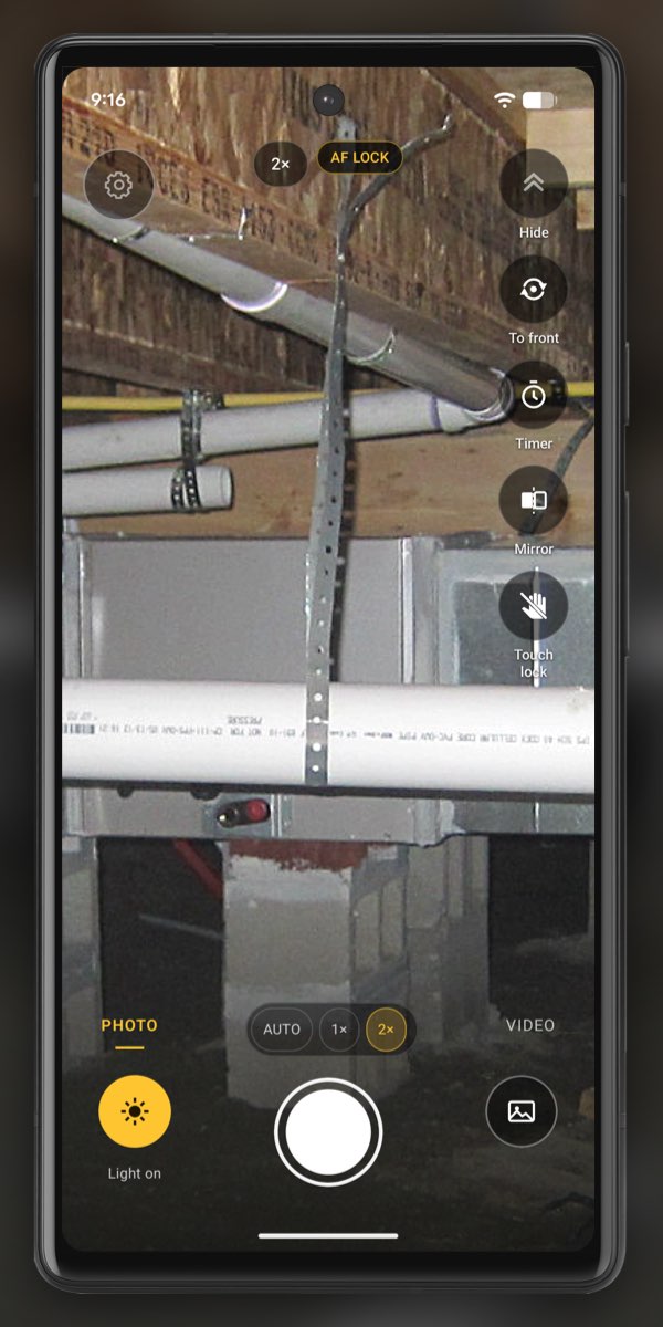 InspectCam zoomed to 2x on white drain pipes and yellow cable under the floor joists of a crawl space, with the 2x and focus lock chips at the top and Light on lit.