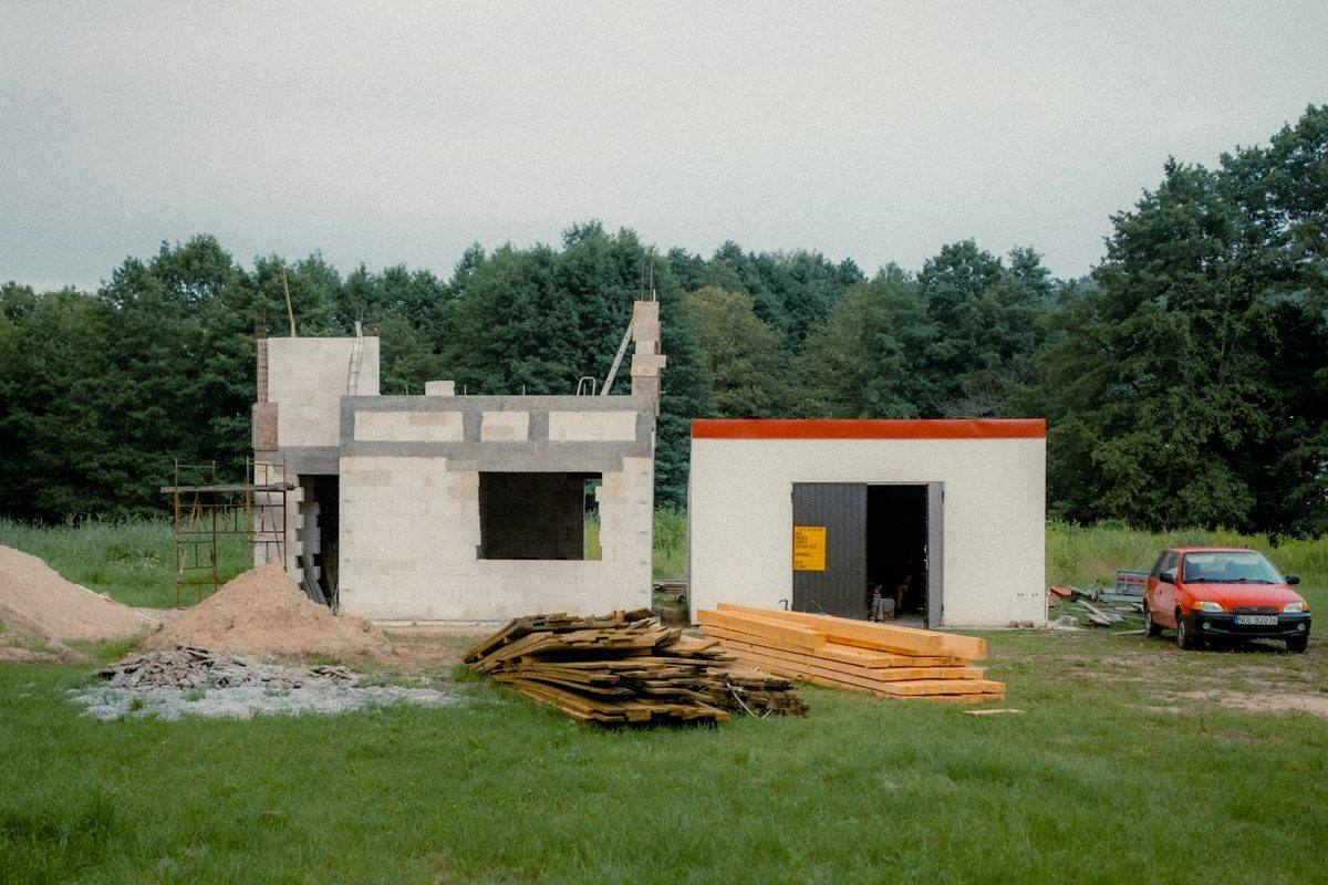 A small block house under construction on a grass lot, with stacks of lumber on the ground in front of it.