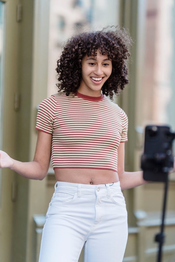 A smiling woman with curly hair talking to a phone on a tripod on a city street.