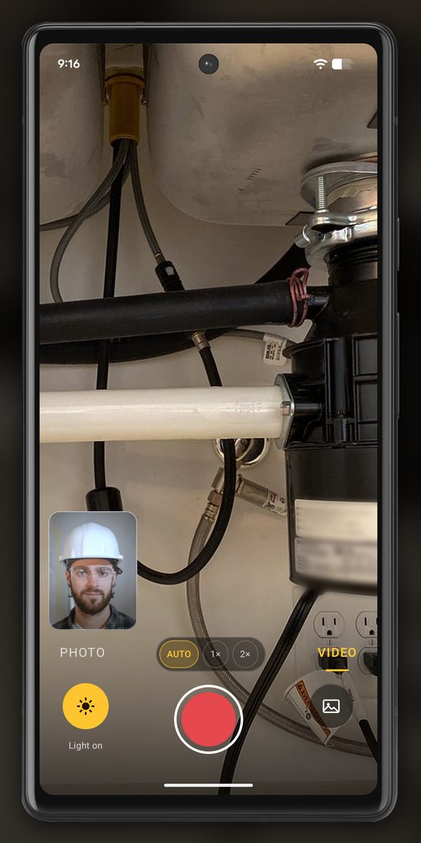 InspectCam’s Dual screen in VIDEO on an Android phone under a kitchen sink: the back camera shows the garbage disposal, the drain pipe and the supply lines with Light on lit, and a small front-camera window at the bottom left shows a worker in a white hard hat and safety glasses.