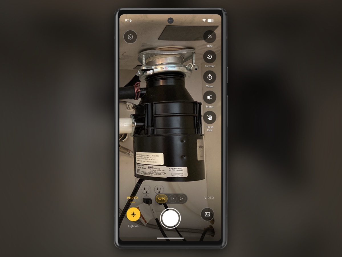 InspectCam open on a phone under a kitchen sink: the garbage disposal and the outlets below it are lit, with the settings gear at the top left, the tool buttons down the right side, and the yellow Light on button and the shutter at the bottom of the screen.