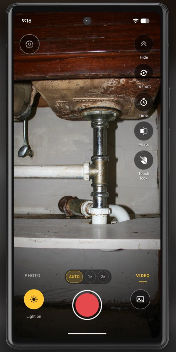 InspectCam in video mode on a phone held into the cabinet under an old kitchen sink: the bottom of the sink, the chrome drain pipe and its tee, the trap going down through a white shelf, and the floor of the cabinet below it, all lit by the phone’s light. The tool buttons run down the right side, with the red record button and the yellow Light on button at the bottom.