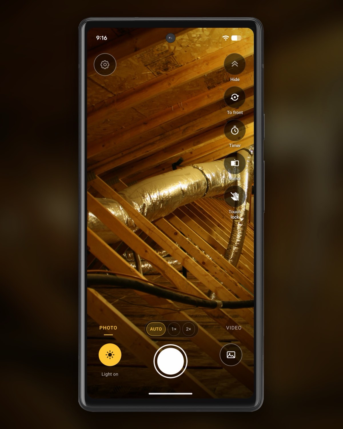 InspectCam’s free camera on an Android phone, held up in an attic: foil-wrapped flex ducts running between the roof trusses, the truss webs and the roof deck lit warm in the middle and fading into the dark at the edges. PHOTO is selected, the yellow Light on button is lit, with the lens chips, the shutter and the gallery button, the settings gear at the top left and the tool buttons down the right, Timer and Touch lock among them.