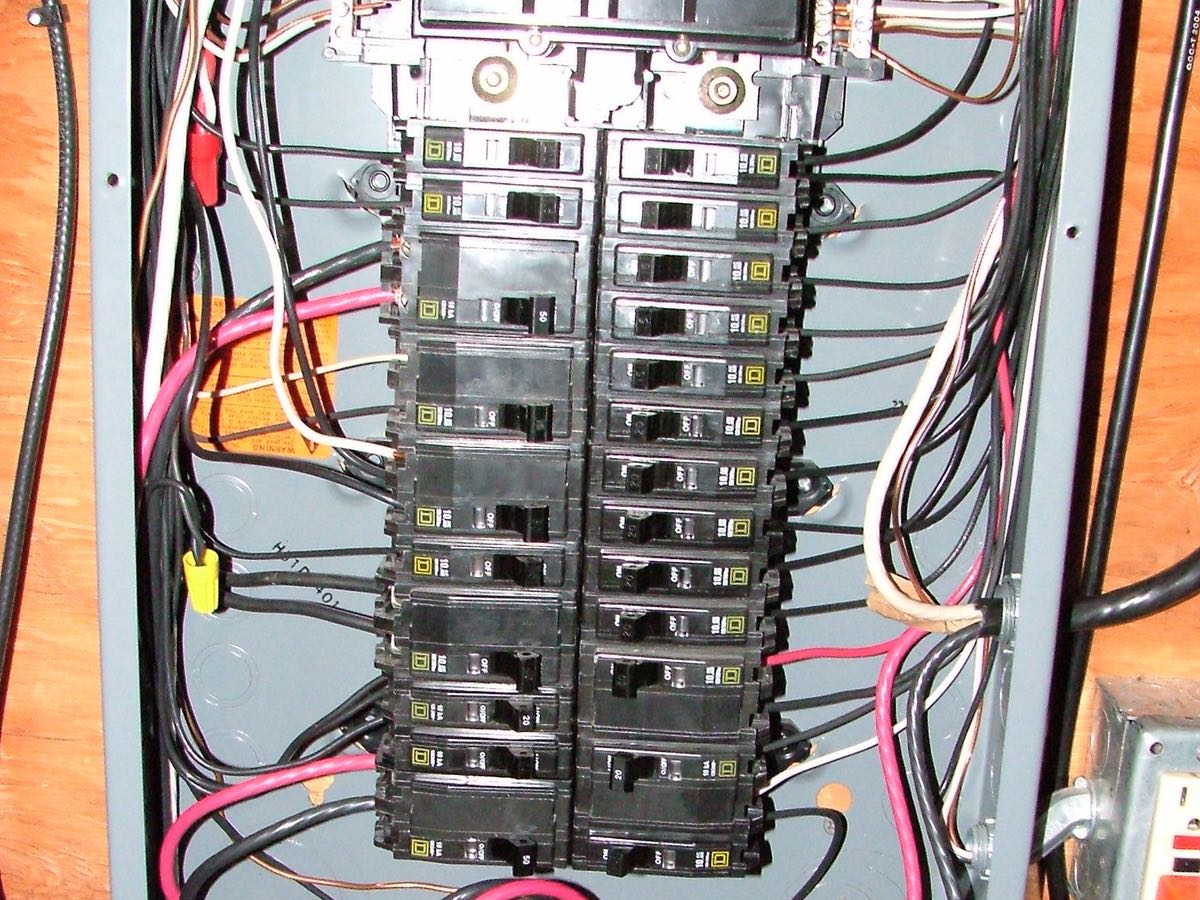 A home electrical panel with its cover off: two columns of black breakers, with black, white, red and bare wires running to them inside a gray steel box on a plywood wall.