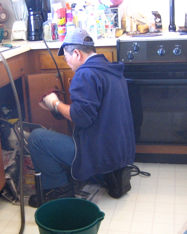 A plumber in a cap and gloves kneeling in front of an open kitchen sink cabinet, working on the pipes inside, with a bucket on the floor beside him.