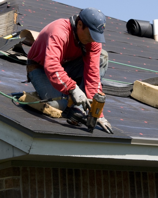 A roofer in a red shirt and gloves kneeling on underlayment at the roof edge with a nail gun.