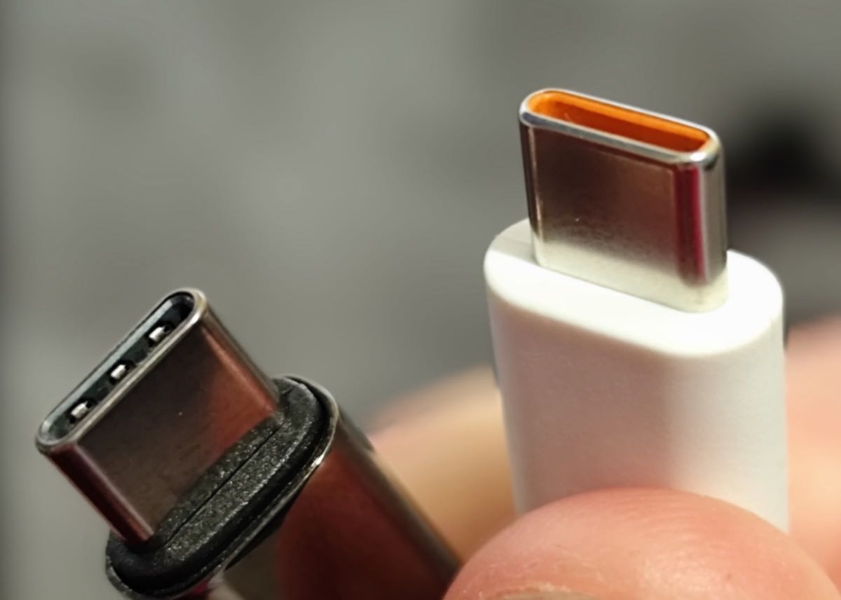 Close-up of two USB-C cable plugs held between finger and thumb, one in a dark metal housing and one in white plastic.