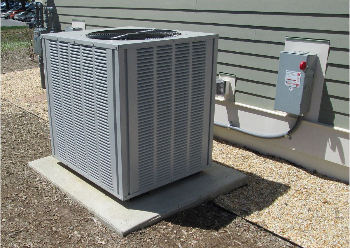 A grey outdoor air conditioning unit on a concrete pad in bright sun, beside a building with a grey disconnect box on the wall.
