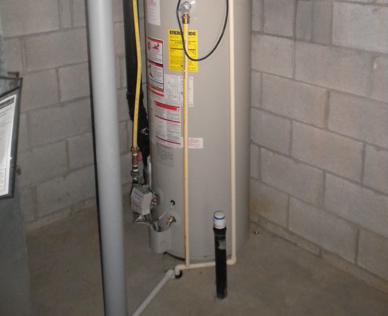 A gas tank water heater in the corner of a basement, its jacket covered in stickers and labels, with gas and water pipes around it.