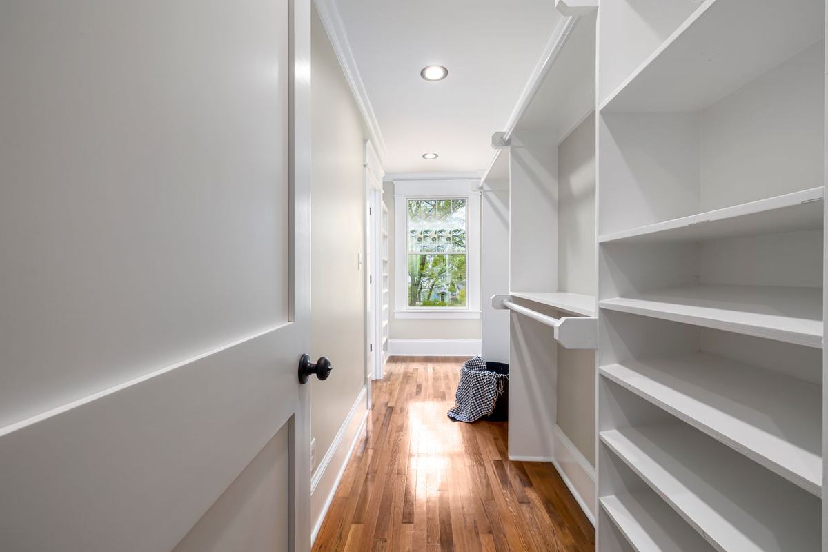 An empty walk-in closet with white shelves, a hanging rod, a wood floor and a window at the end.
