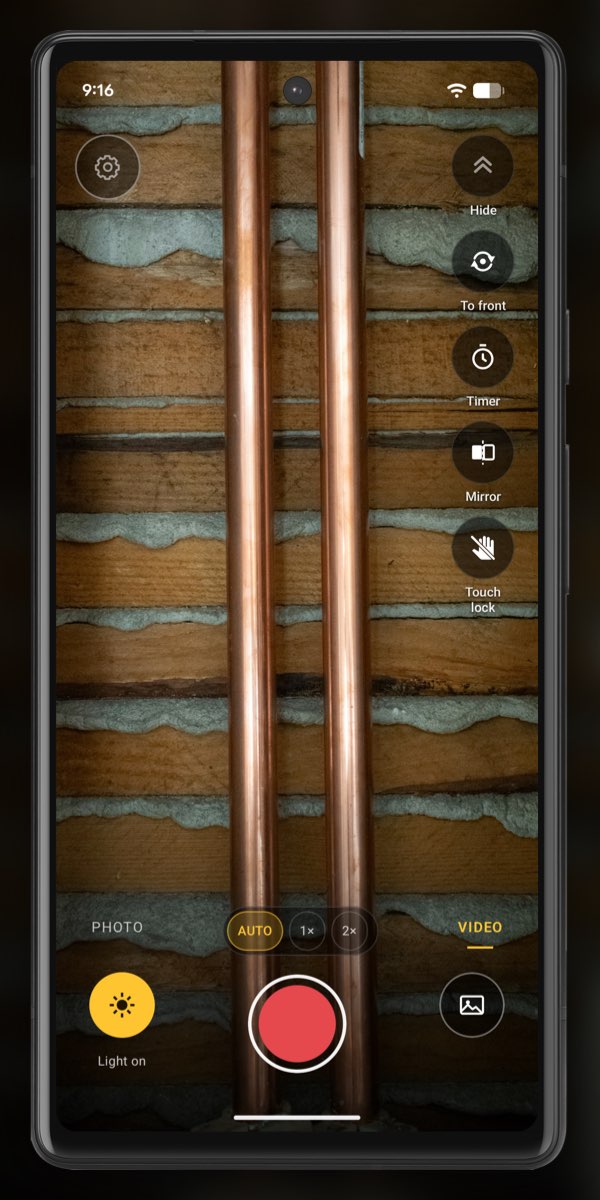 InspectCam in VIDEO mode on an Android phone, aimed at two copper pipes in an opened wall, with bright streaks of reflected light running down each pipe. Light on is lit at the bottom left and the red record button is in the middle.