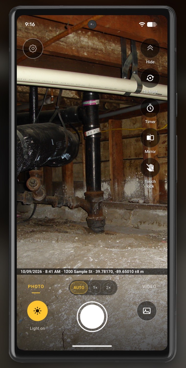 The camera on a phone in a crawl space, pointed at a black drain stack with a rusted fitting at its foot, ducts and pipes overhead and white staining on the wood wall behind. Just above the Photo, zoom and Video row runs one thin dark line of small white text: the stamp line, with a date, a time, a sample street address and GPS coordinates.