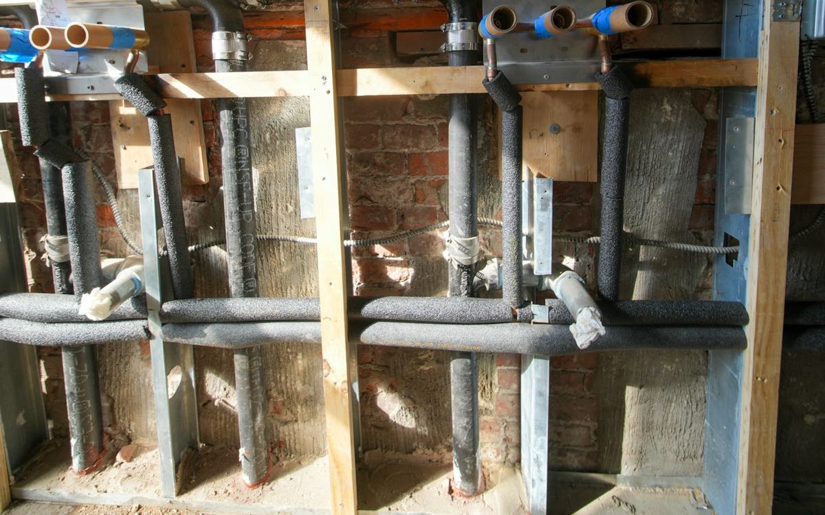 An opened-up wall with copper and plastic water pipes and electrical conduit running through the stud bays.
