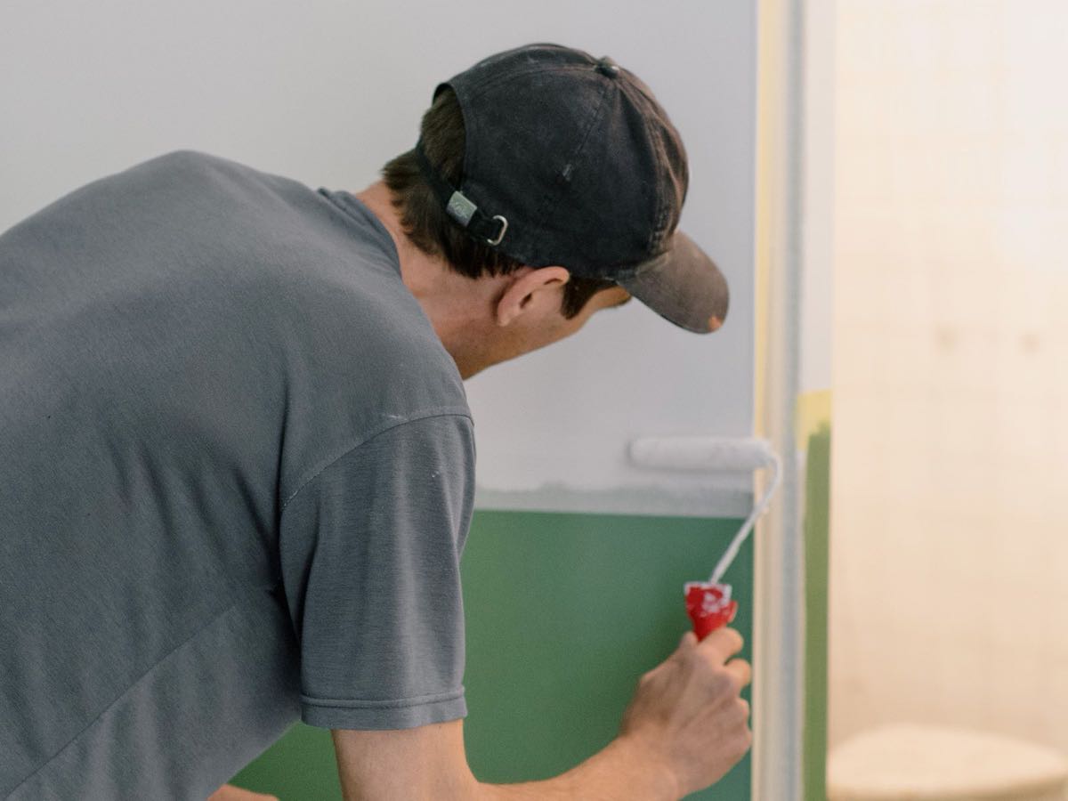 A painter in a cap and gray T-shirt rolls white paint over a green wall in an empty room.