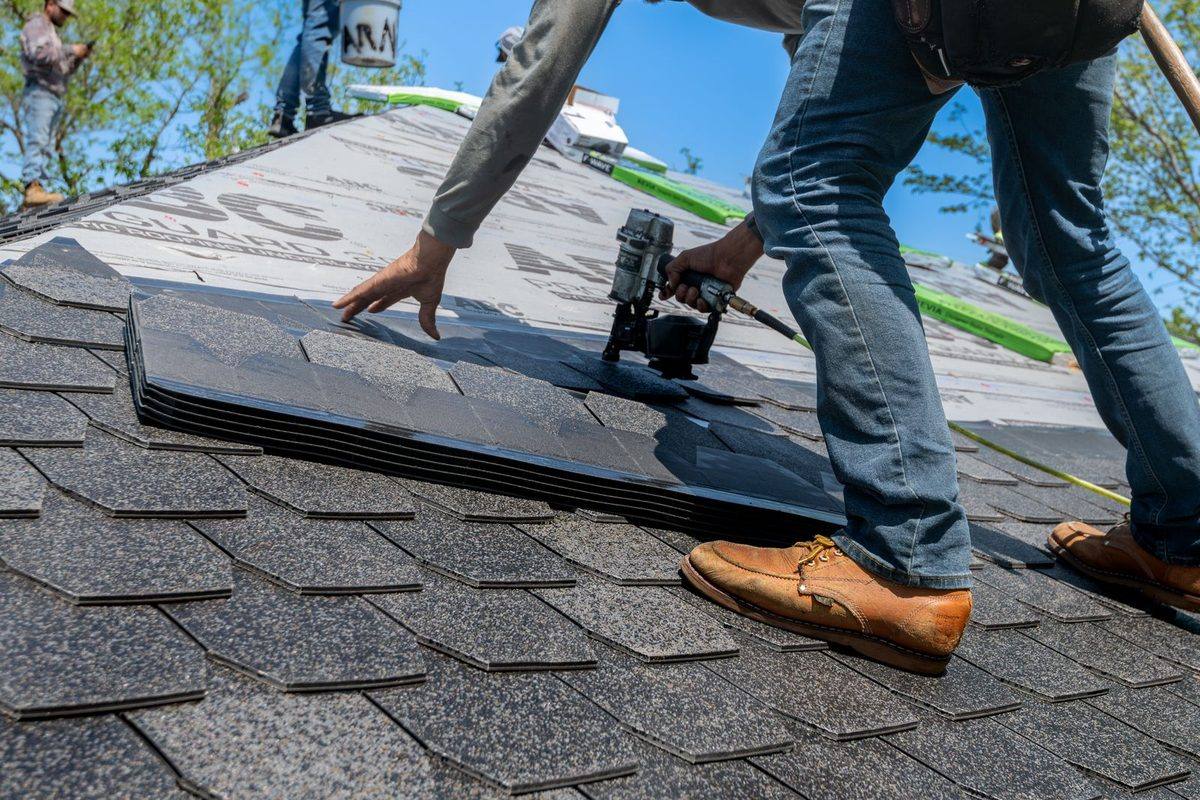 A roofer kneeling on a roof nailing new dark gray shingles with a nail gun, under a blue sky.