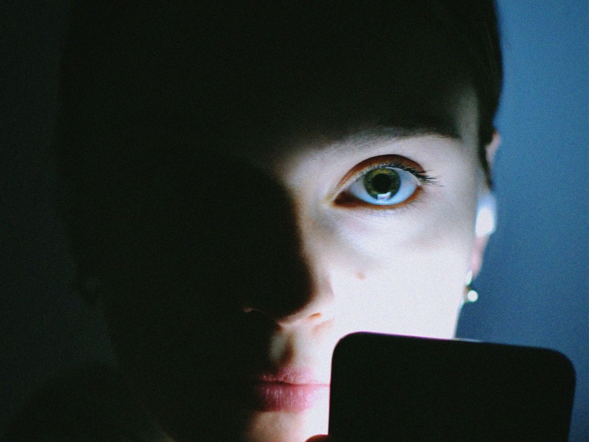A woman in a dark room holding a phone just below her chin; its light catches one side of her face and leaves the other in shadow.