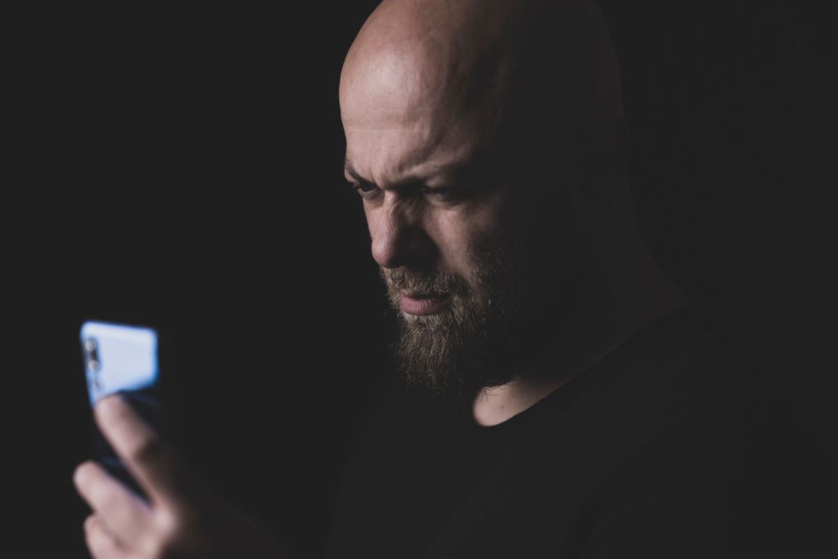 A bald, bearded man in a dark room frowning at the phone in his hand.