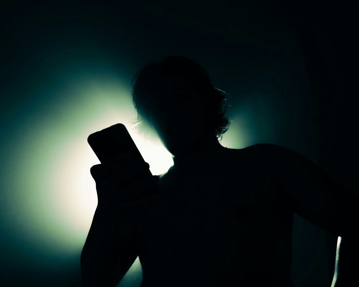 A person in a dark room holding up a phone, lit only from behind, so the face is a dark silhouette.