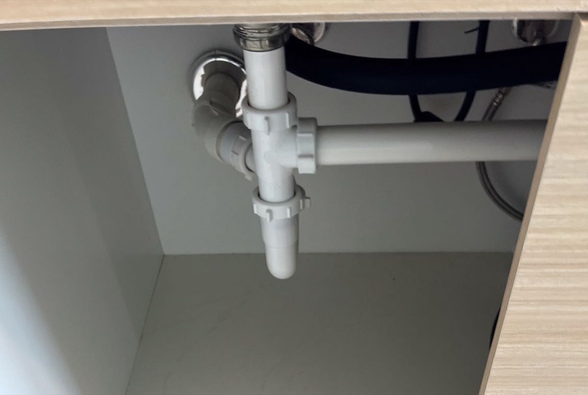 An emptied sink cabinet: white plastic drain pipes with a slip nut at every joint, a black hose and a braided supply line at the back.