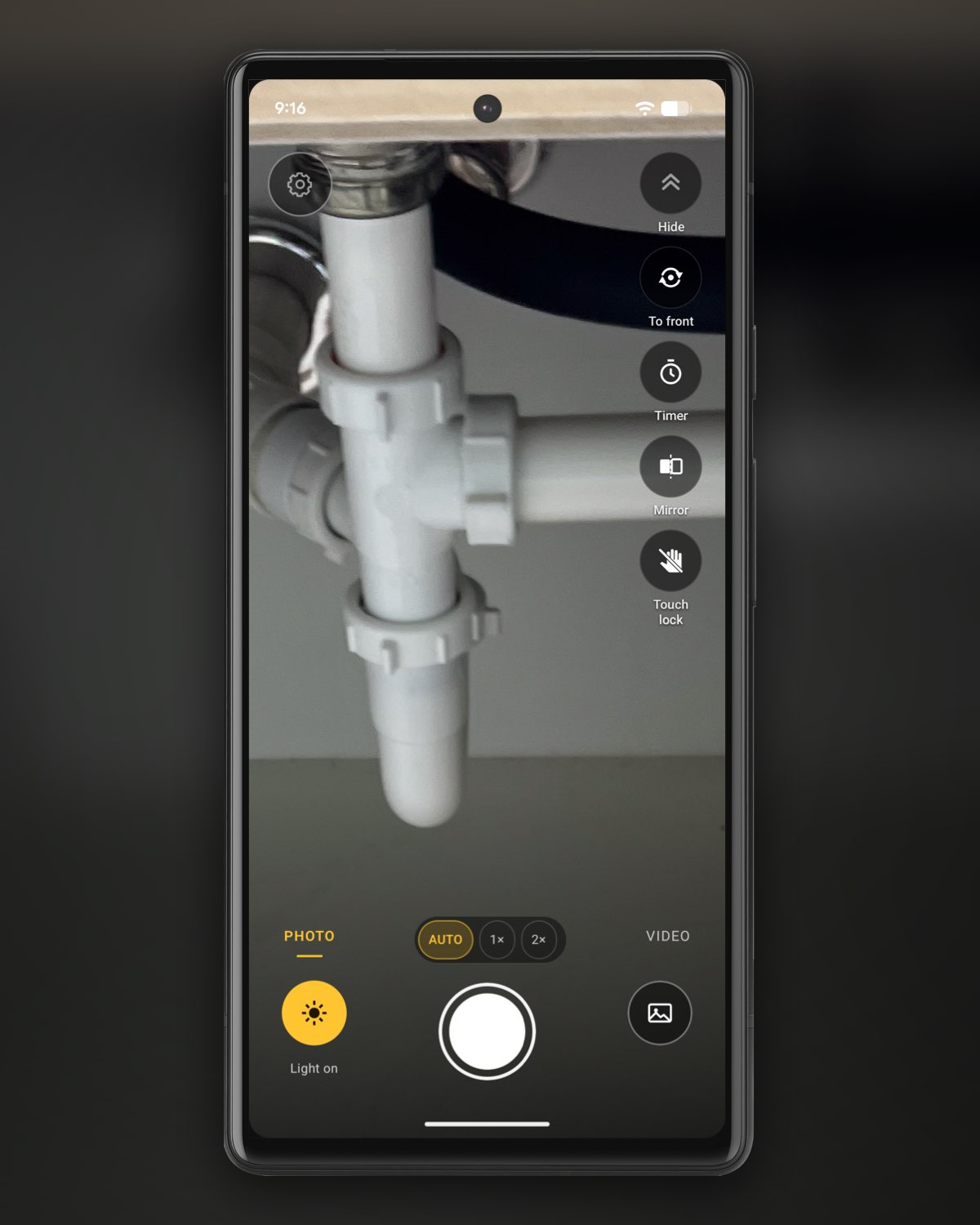 Illustration: a phone running InspectCam’s camera under a sink, the white plastic drain and its slip nuts in view, with the yellow Light on button lit and Photo selected, the settings gear at the top left and the Hide, To front, Timer, Mirror and Touch lock buttons down the right side.
