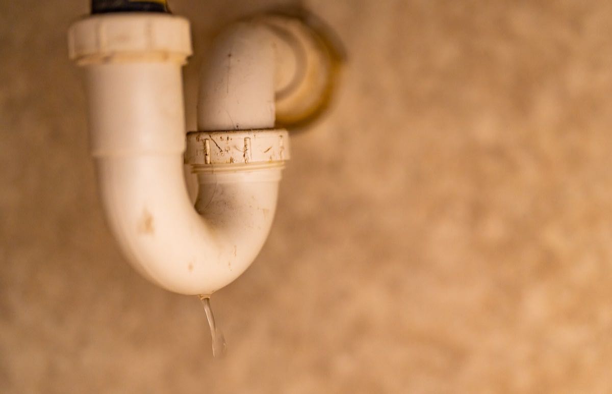 A white plastic P-trap under a sink with a drop of water hanging from the slip nut.