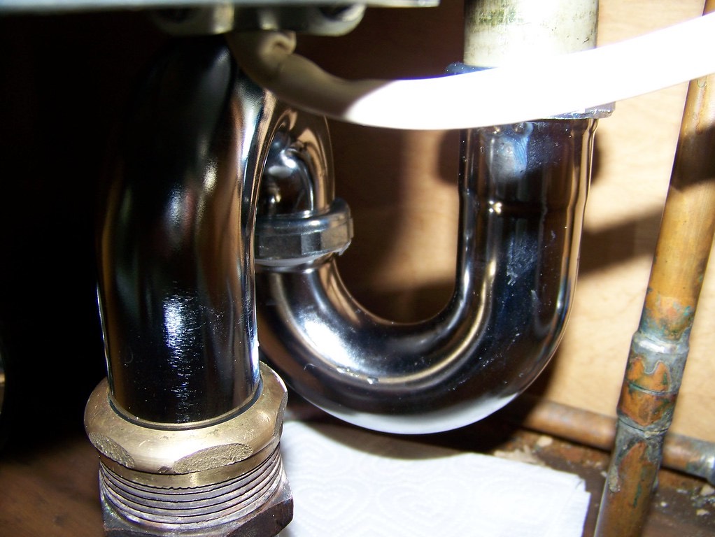 A chrome trap under a sink, seen close up: a nut where the curved trap joins the pipe coming down from the sink, a brass nut on the pipe at the left, and a paper towel laid flat underneath.