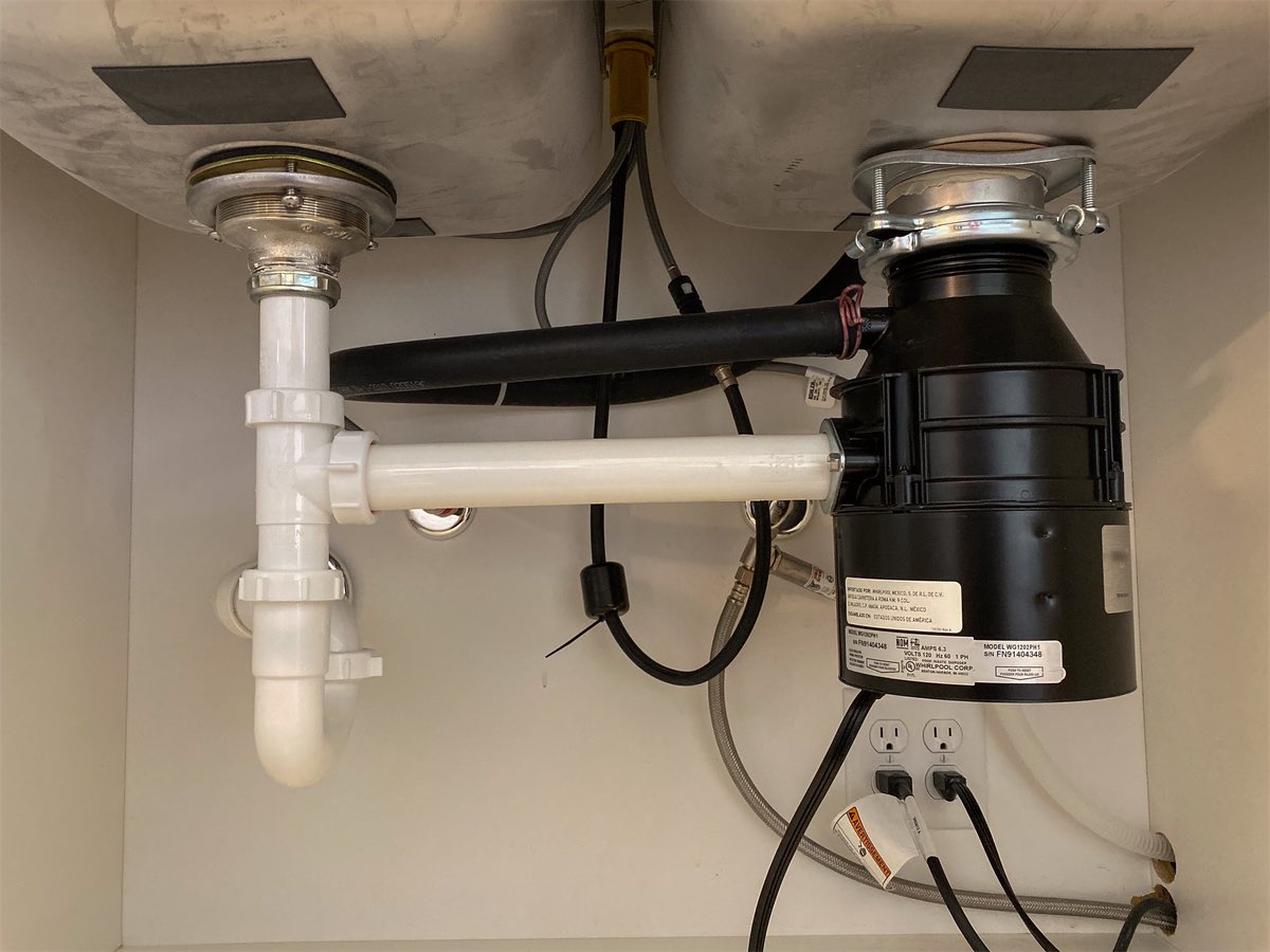 The plumbing under a double kitchen sink: a white plastic drain with slip nuts and a trap on the left, a garbage disposal on the right with a black dishwasher hose running into it, and braided supply lines in the middle.
