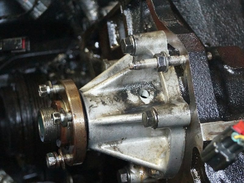 Close view deep in an engine bay: a pulley, a drive belt and a metal housing with bolts and fluid residue.