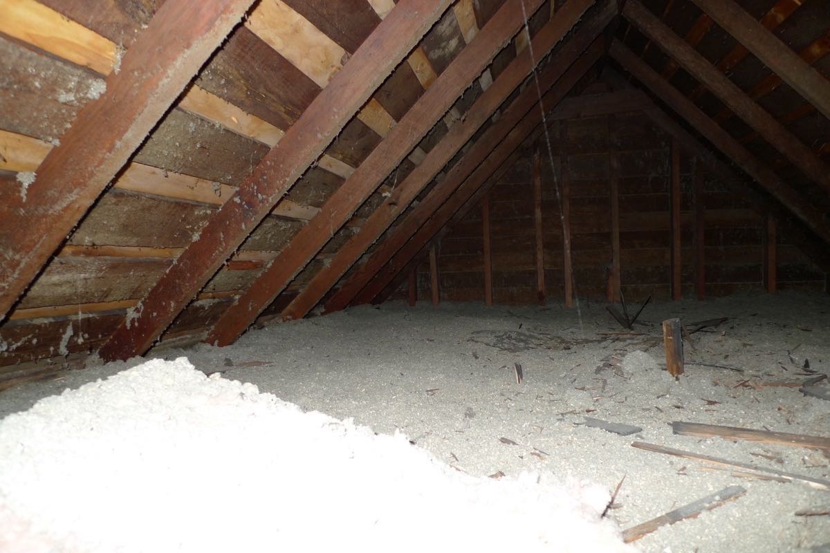 An unfinished attic seen from near the hatch: rafters on board sheathing, blown-in insulation across the floor and a gable end at the back.