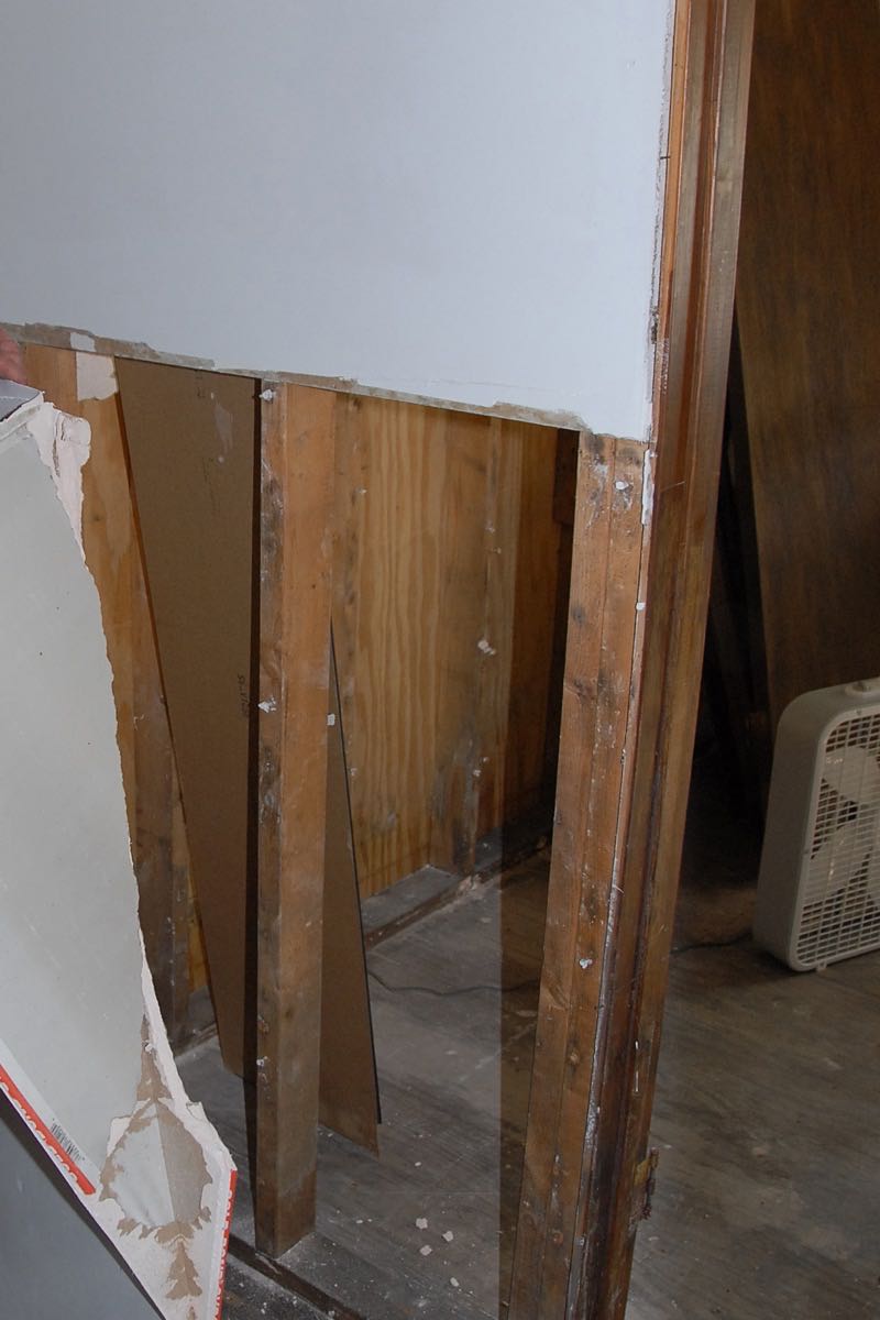 A wall with its lower drywall cut away in a straight line, showing the wood studs and paneling behind it, the removed piece of drywall at the edge of the frame and a box fan on the floor beyond.