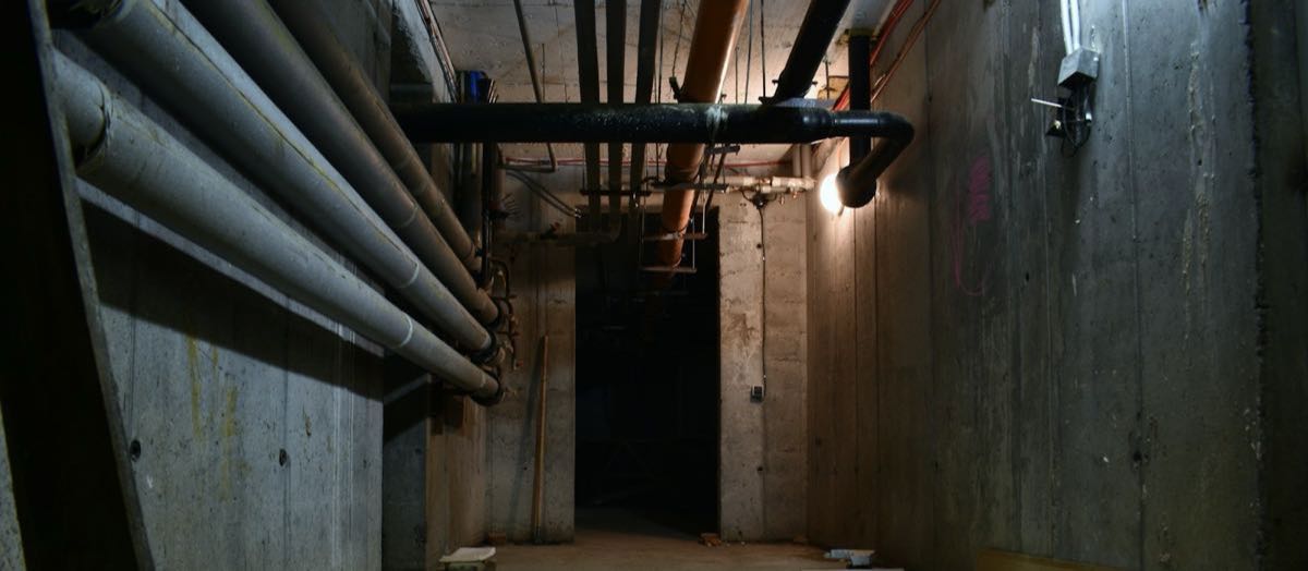 A dim concrete service corridor with metal pipes along the walls and ceiling.