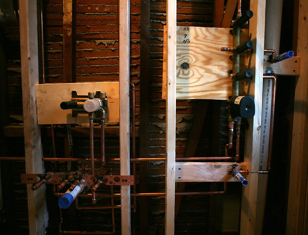 The inside of an opened bathroom wall: wood studs, copper supply pipes and shower valves on wood blocking, capped pipe ends, and old brick and plaster behind.