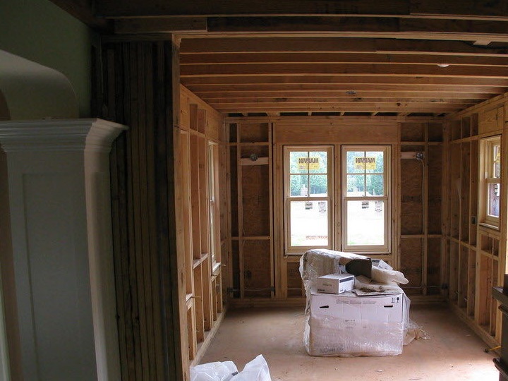 A room addition at the framing stage, seen from the house: bare studs and sheathing on every wall, two new windows, open ceiling joists overhead and boxed materials on the subfloor.