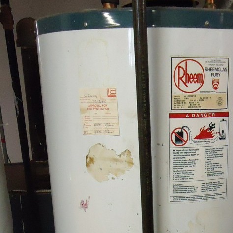 A gas water heater in a basement, its jacket carrying a rating plate, a danger label and a fire-protection approval tag, with a gas pipe running down its front and old pipes along the wall behind.